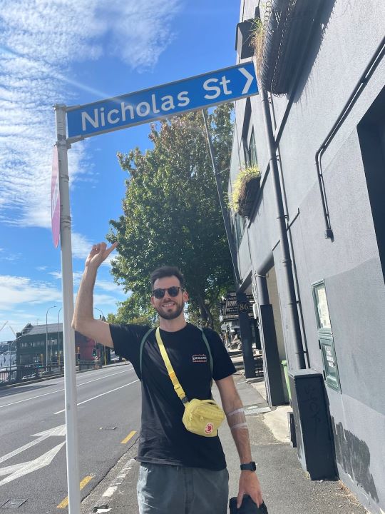 "Ich stehe unter einem blauen, leicht bewölkten Himmel und zeige lächelnd auf ein blaues Straßenschild auf dem "Nicholas Street" steht. Ich trage ein schwarzes T-Shirt, eine graue Hose, eine gelbe Bauchtasche und eine Sonnenbrille."