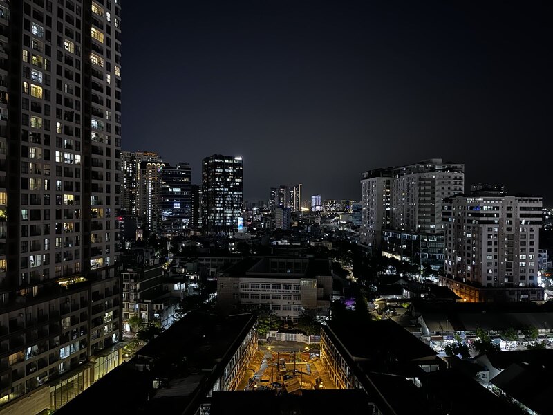 "Ein Blick über Ho Chi Minh City bei Nacht. Neben einem Hochhaus sind einige kleinere Gebäude mit flachen Dächern. In einigen Fenstern ist das Licht an. Im Hintergrund erkennt man einige Hochhäuser bei dunklem Himmel."