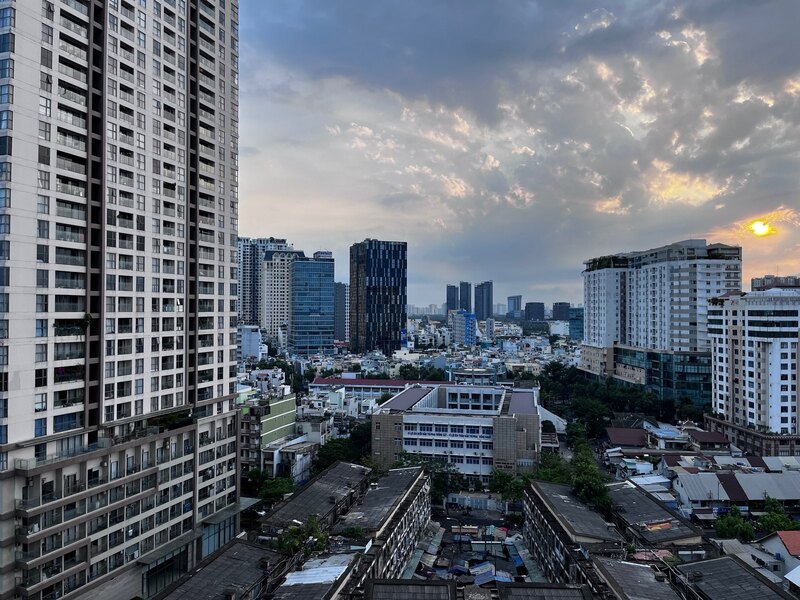 "Ein Blick über Ho Chi Minh City. Neben einem Hochhaus sind einige kleinere Gebäude mit flachen Dächern. Im Hintergrund erkennt man einige Hochhäuser, einen wolkenverhangenen Himmel und die aufgehende Sonne"