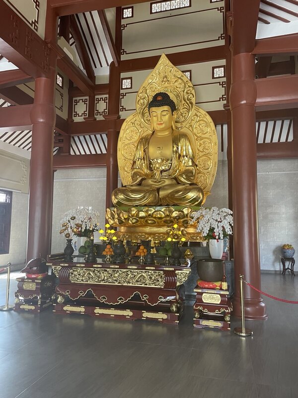 "Eine goldene Buddhastatue steht auf einem Podest. Davor ist ein Holztisch auf dem mehrere Zitrusfrüchte in mehreren Obstschalen stehen."