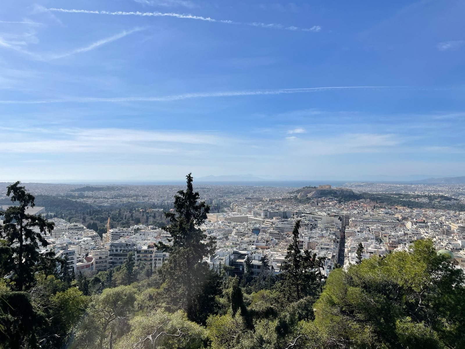 Der Blick vom Lykabettus Richtung Akropolis. Ein blauer Himmel mit leichtem Wolkenschleier und Kondensstreifen. Darunter weiße Häuser und Grünflächen, in der Mitte auf einer Erhebung, die Akropolis.