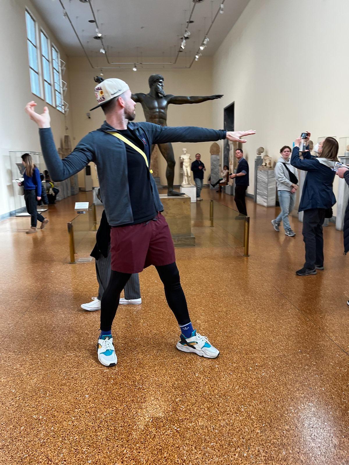 Ich stehe auf dem hellorangen Boden des Museums vor einer dunklen, brozenen Statue. Ich trage eine nach hinten gedrehte, beige Kappe, eine dunkelgraue Fleecejacke, eine rote, kurze Hose mit Leggins und weiße Sneaker. Ich ahme die Geste der Statue nach: Ich stehe seitlich, den rechten Arm habe ich abgewinkelt nach oben und den linken Arm nach vorne gerichtet.