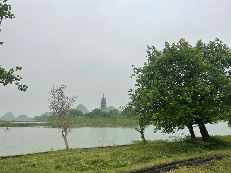 "Eine Pagode steht auf einer kleinen grünen Insel des Sees. Dahinter ragen abgerundete Berge in die Höhe."