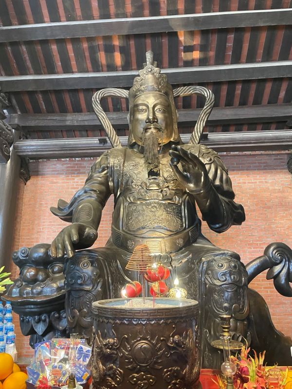 "Eine Wächterstatue aus Kupfer mit freundlichem Blick. Sie trägt eine Rüstung und einen hält in ihrer Hand eine Perle in die Höhe. Vor ihr befindet sich ein Räuchergefäß und Opfergaben. "