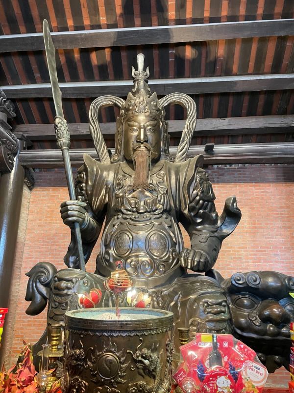"Eine Wächterstatue aus Kupfer mit grimmigem Blick. Sie trägt eine Rüstung und einen Speer. Vor ihr befindet sich ein Räuchergefäß und Opfergaben. "
