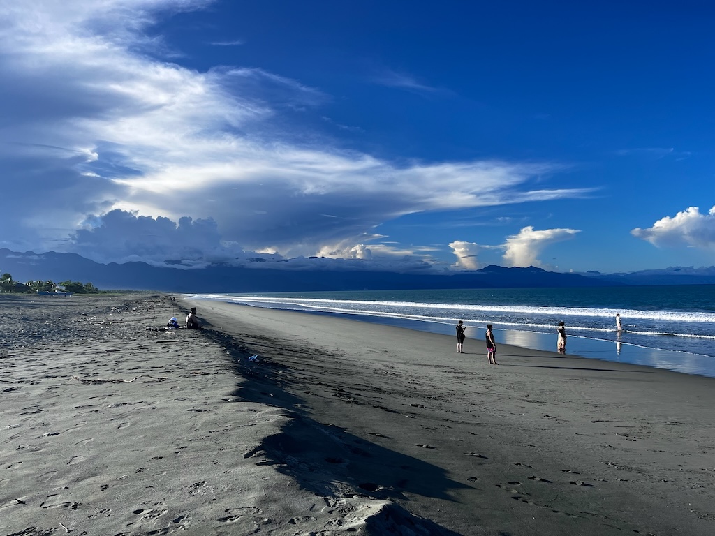 "Ein grauer Sandstrand, der eine kleine Düne bildet. Darauf sitzt ein Mann. Davor stehen einige Leute am Strand. In der Ferne sieht man ein dunkelblaues Gebirge. Darüber türmen sich die Wolken auf. Der Himmel ist blau."