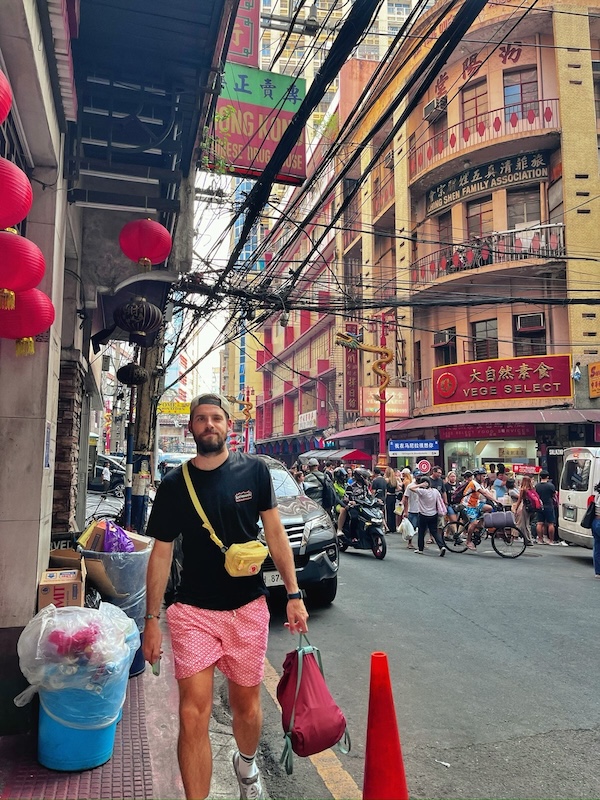 "Ich spaziere der Kamera entgegen. Hinter mir sind einige Geschäfte mit chinesischen Schriftzeichen, einige Menschen zu Fuß und ein Mopedfahrer."