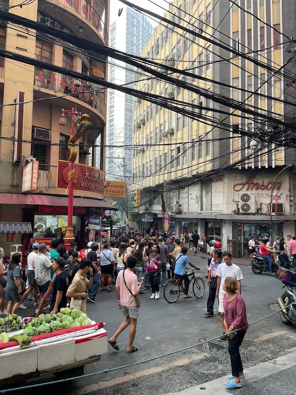 "Eine Straßenszene in Binondo. Viele Menschen sind zu Fuß unterwegs und bummeln über den Markt. Die verworrene Stromverkabelung sticht ins Auge."
