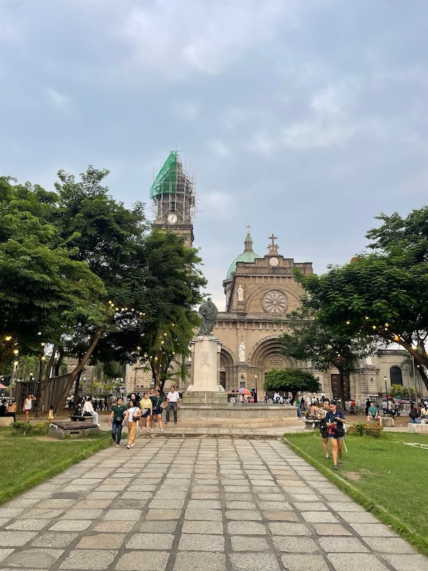 "Eine Kathedrale hinter einem grünen Park. Einige Menschen sind auf dem vorliegenden Platz unterwegs."