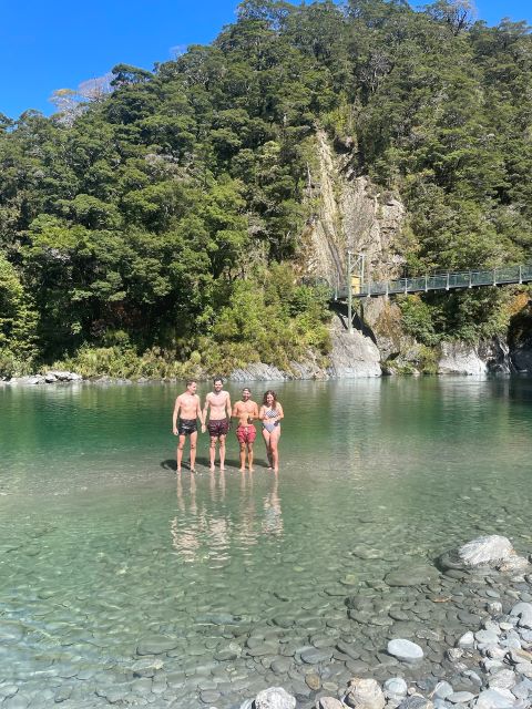 "Ein grünes Flussbecken unter einer Hängebrücke. Im Wasser stehen Manu, ich, Simon und Sophia in Badeanzug."