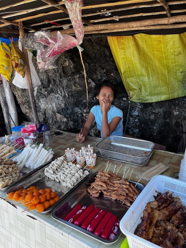 "Ein junges Mädchen mit blauem T-Shirt sitzt am Thresen ihres Essenstands. Hier gibt es verschiedenes Essen auf Spießen, das frittiert wird. "