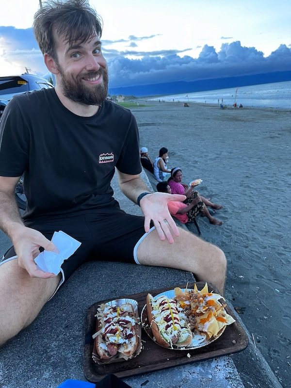 "Ich sitze lächeln an der Promenade und deute auf das Essenstablett vor mir, auf dem zwei Hotdogs mit sehr viel Sauce stehen"