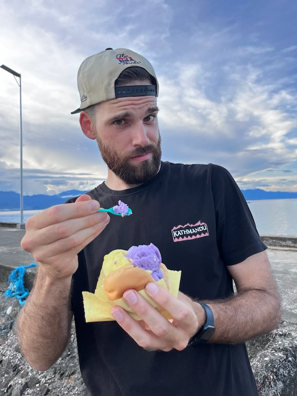 "Mit fragendem Blick halte ich meinen kleinen Plastiklöffel in die Kamera, auf dem etwas Ube-Eis ist, welches ich in meiner anderen Hand halte."