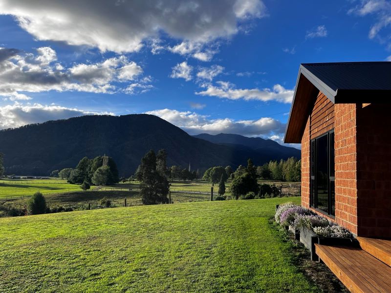 "Vom Garten der Unterkunft aus sieht man dunkelblaue Berge und einen bewölkten Himmel. Die Sonne scheint auf den grünen Rasen und das Haus mit Backsteinziegeln."