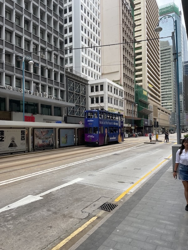 "Auf der Straße, umgeben von riesigen Hochhäusern fährt eine blau-violette Doppeldecker-Tram."