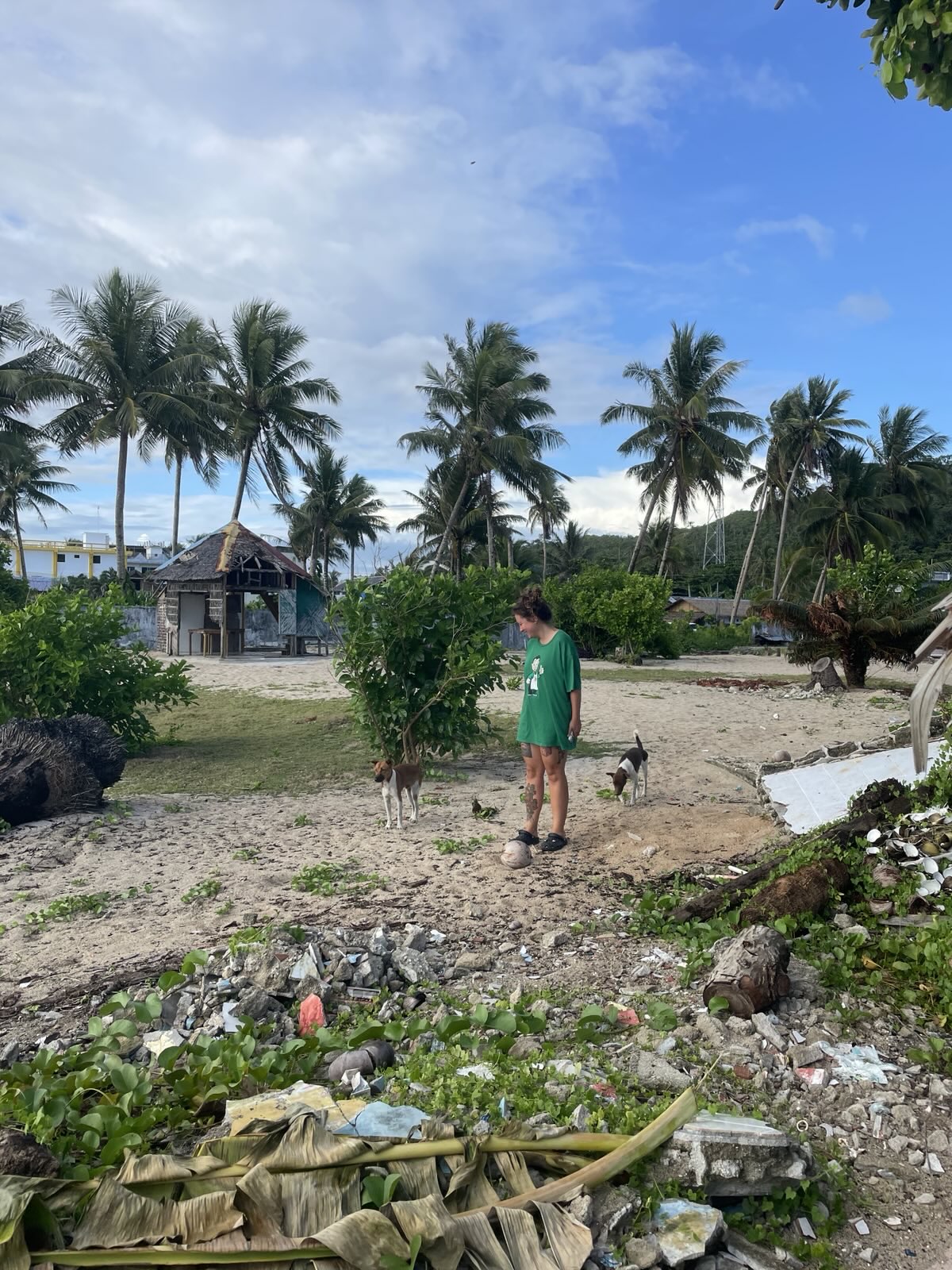 "Sophia steht vor einigen grünen Palmen, Büschen und einer Holzhütte mit Strohdach. Neben ihr sind zwei Hunde."