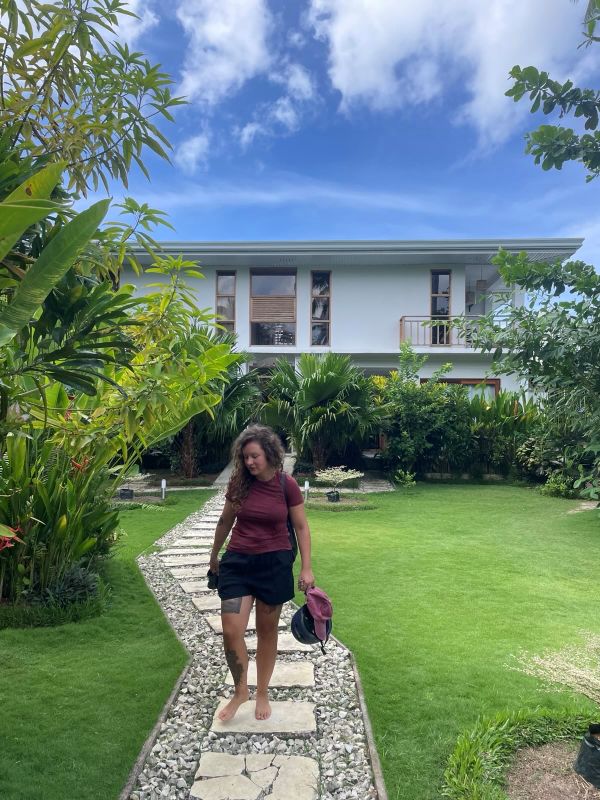 "Sophia spaziert den mit Stein gepflasterten Weg entlang, der durch den grünen Garten führt. Sie trägt ein rotes Shirt und eine kurze schwarze Hose."