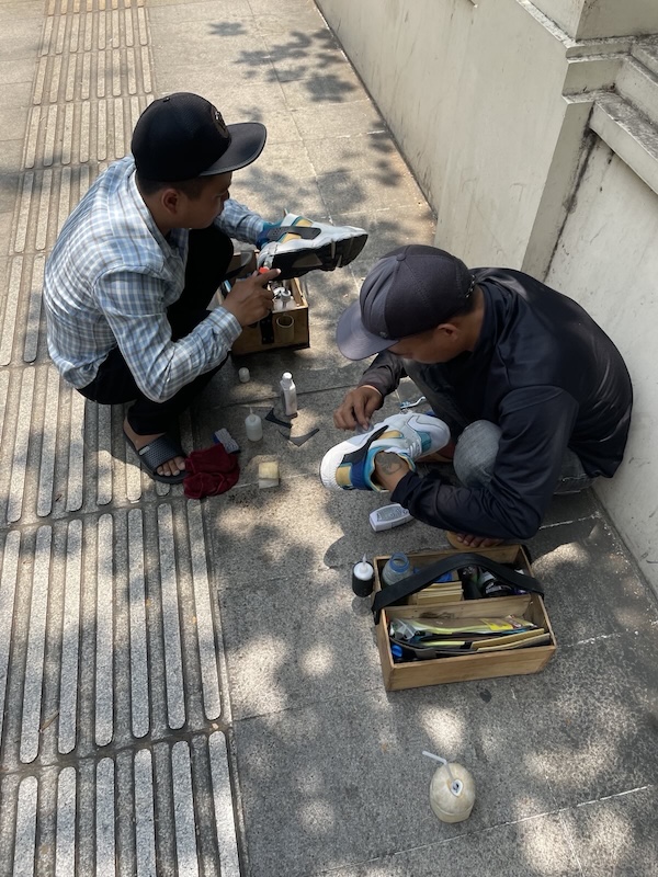 "Zwei Straßenhändler stehen gebücht auf der Straße und arbeiten an meinen Schuhen. Der eine schneidet mit Stanley-Messer eine Sohle zurecht, der andere flickt kleine Löcher."