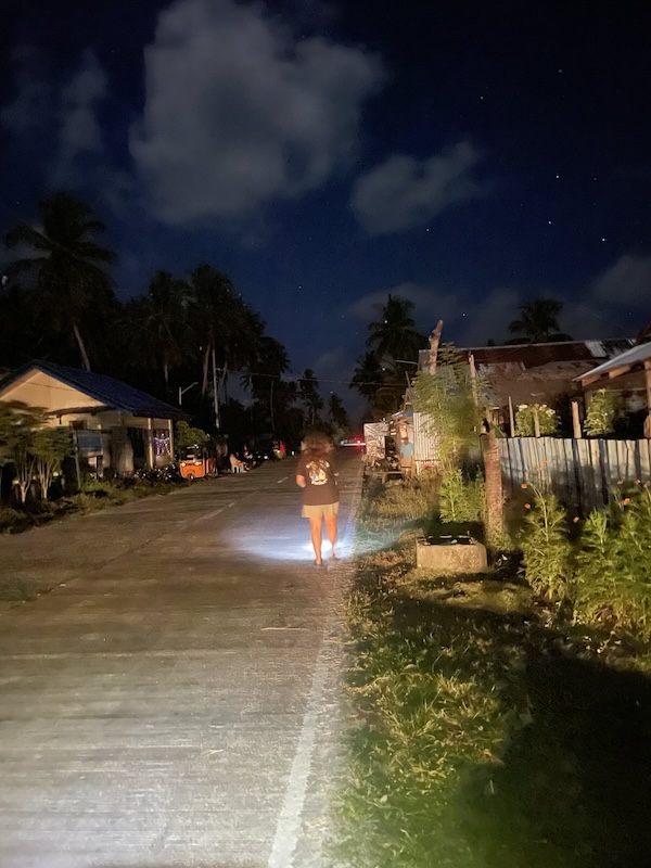 "Sophia spaziert mit Taschenlampe am Rande der dunklen Straße entlang. In der Ferne erkennt man das Rücklicht eines Mopeds. Die Straße wird von den Scheinwerfern eines Autos ausgestrahlt."