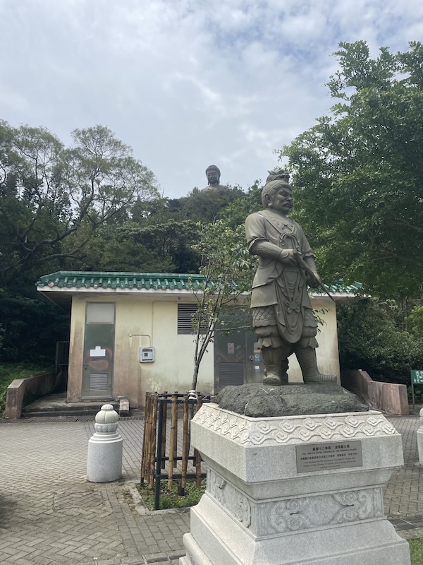 "Eine Steinstatue eines Generals, der einen Bogen in der Hand hält. Auf seinem Kopf trägt er einen Drachen. Hinter dem General im Grünen ist der große Buddha."
