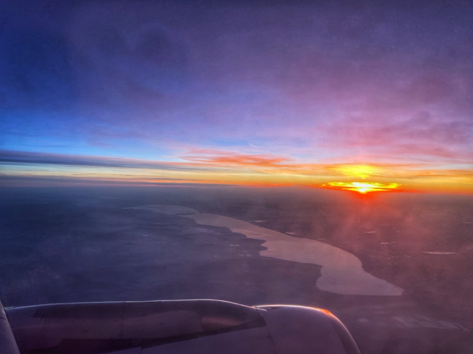 Der Sonnenaufgang vom Flugzeug aus wirft den dunkelblauen Himmel in ein Licht das vom Gelben, über's Orange bis hin zu rot geht. Unter der Sonne ist der ungarische See Balaton zu sehen.