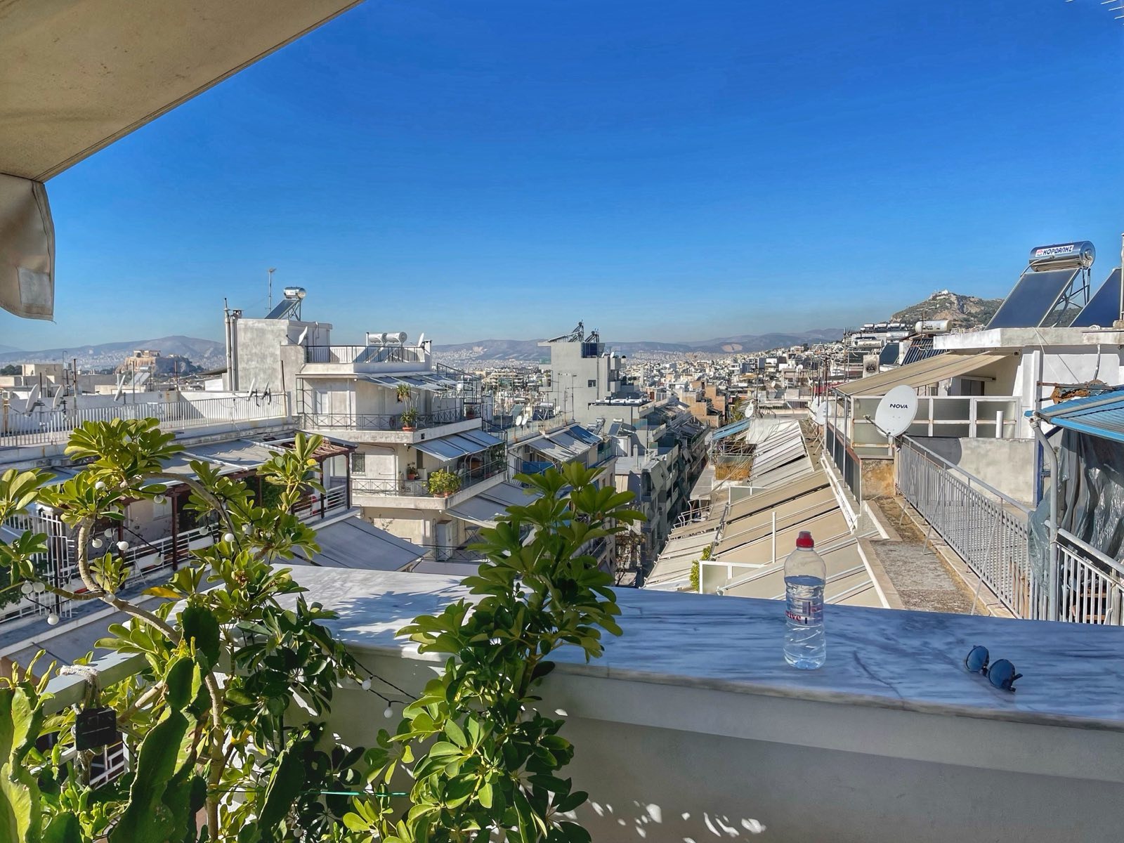 Blauer Himmel über der Terrasse, auf dessen Vorsprung eine Mamorplatte als Ablage dient. Darauf steht eine Plastikflasche und eine Sonnenbrille. In der Ferne sieht man das Stadtbild Athens: weiße Häuser, die Akropolis zur Linken und der Stadtberg Lykabettus zur Rechten.