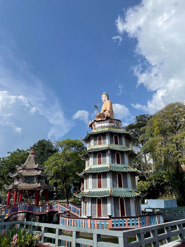 Auf einem mehrstöckigen Tempel mit grünen Dächern steht eine Buddha-Figur mit oranger Robe und türkiser Kopfbedeckung. Der Tempel ist umgeben von grün blühenden Bäumen und einer Brücke zu einem zweiten Tempel mit roten Säulen und braunem Dach.