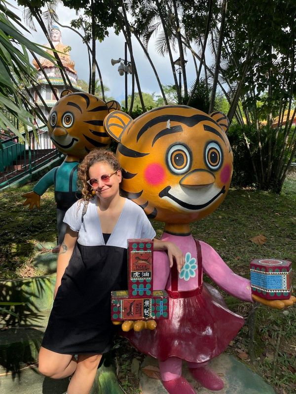 Sophia lehnt sich an eine Tigerstatue die auf zwei Beinen steht und ein rosa Kleid trägt. Die Statue hat ein cartoon-artiges, lächelndes Gesicht und rot angelaufenen Wangen. Sie hält in beiden Hände Tiger Balm Produkte. Sophia lächelt. Sie trägt ihre Haare zusammengebunden, hat eine Sonnebrille auf und trägt ein blau-schwarzes Kleid.