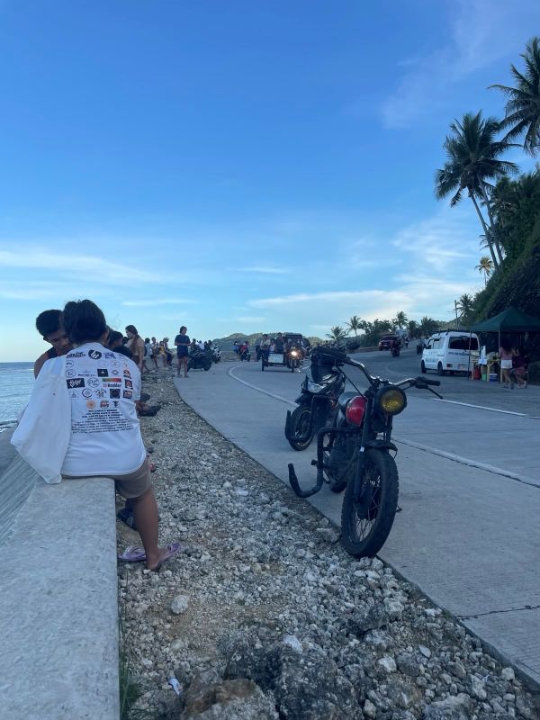 "Am Straßenrand gibt es eine Sitzfläche, auf der einige Menschen sitzen. Hier parken auch einige Mopeds. Ein Tuktuk befährt die Straße."