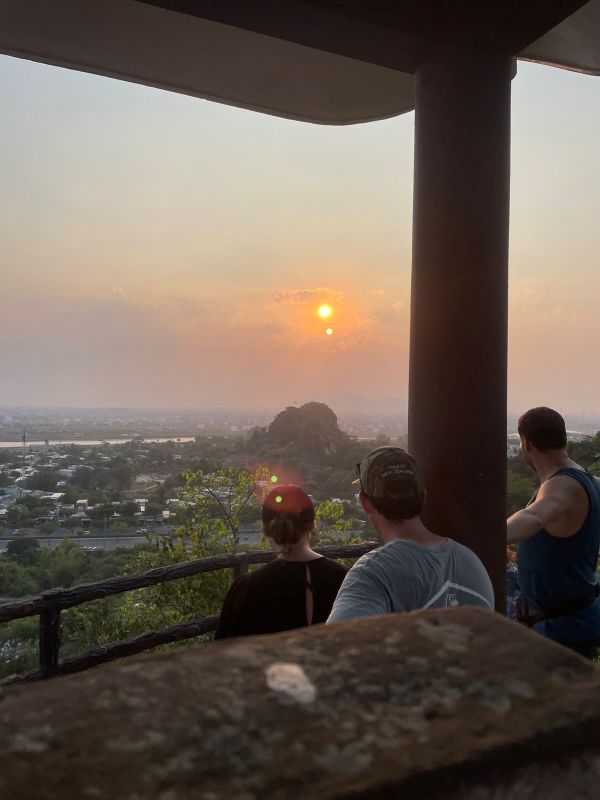 "Die Sonne steht tief über einer Landschaft voller Häuser und einem grünen Hügel. Drei Personen schauen sich den Sonnenuntergang hinter einem Geländer an."