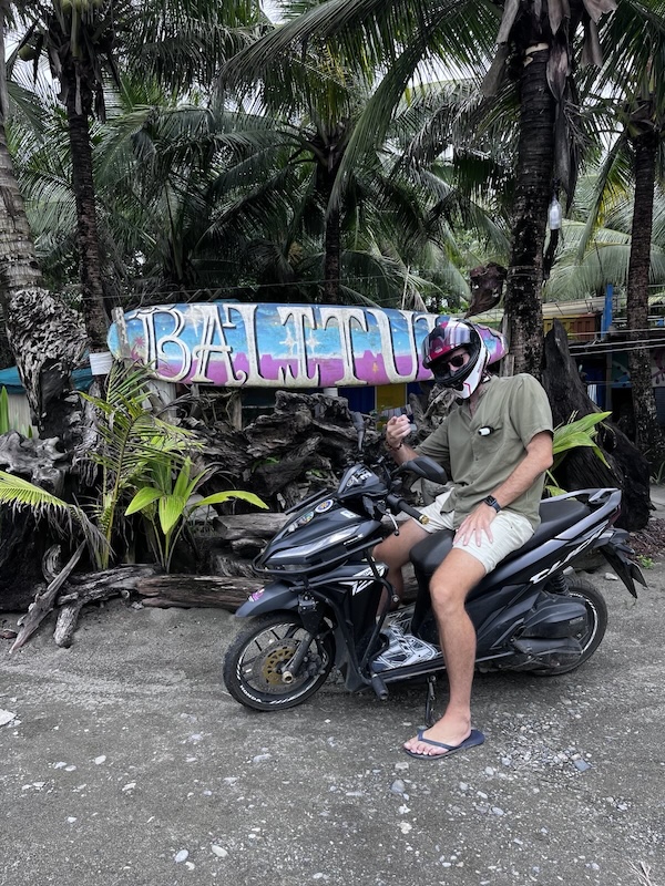"Ich posiere vor einem am Baum befestigten Surfbret, auf dem groß Balituk steht. Ich sitze dabei auf Mofinger und zeige mit dem Finger in die Kamera."
