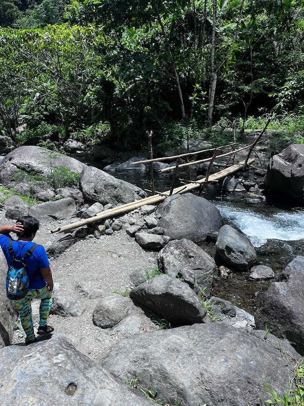 "Sandra trägt ein blaues Shirt und einen Rucksack. Sie geht den Weg entlang. Vor ihr ist eine kleine Brücke, die aus Bambusstöcken gebaut wurde. Darunter fließt ein Fluss."
