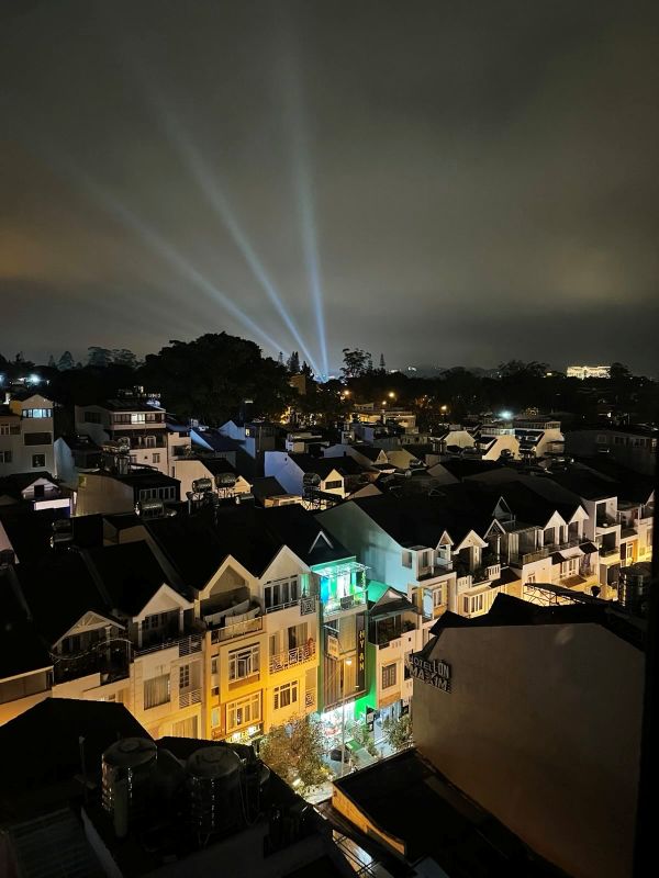 "Der Blick aus dem Hotelfenster zeigt eine hell beleuchtete Straße mit gelbem und grünen Licht. Dahinter strahlen drei Lichtkegel in den bewölkten Himmel."