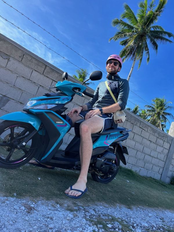 "Ich sitze stolz auf dem blauen Moped namens Mofi und lächle. Ich trage einen rosa Helm und eine graue Weste. Hinter mir ragt eine Palme in die Höhe."