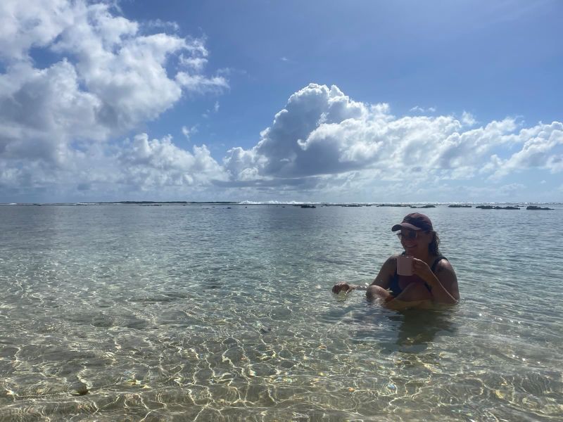 "Sophia sitzt im Bikini im Meerwasser. Sie trägt eine Sonnenbrille und ihre Kappe. In der Hand hält sie eine pinke Tasse Kaffee und lächelt"