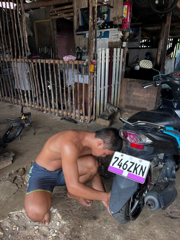 "Eine der leiberllosen Filipinos, wie er am Hinterreifen von Mofi herumwerkelt. Er steht gebückt neben dem Moped und hat den Blick nach unten gerichtet."
