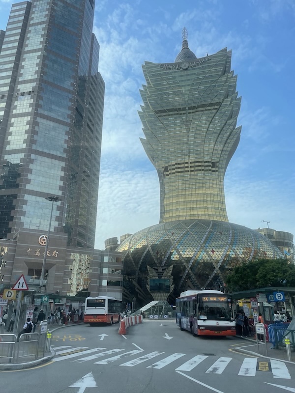 "Ein pompöses Gebäude mit goldenen Fenstern auf dem "Grand Lisboa steht". Darunter ist ein rundes Gebäude und eine Bushaltestelle."