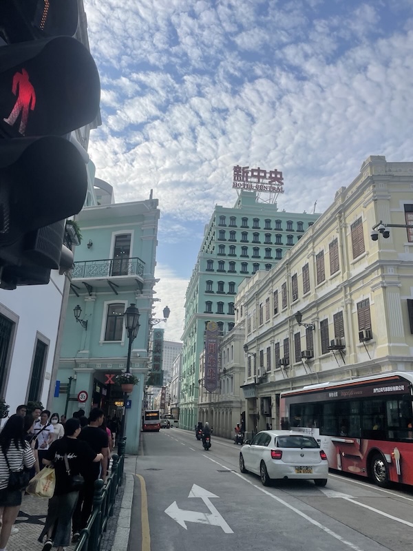 "Eine Straße in Macau zeigt türkisfarbene Gebäude. Eines davon hat ein rotes Schild auf dem "Hotel Central" steht. Darüber ist der Name auf chinesisch abgebildet."