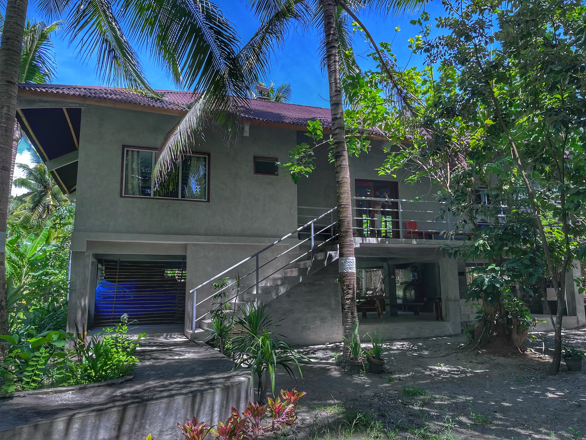 "Unser Haus, vor dem eine große Palme steht. Ein Geländer führt nach oben auf die Terrasse. Rund um das Haus wachsen Büsche und Palmen."