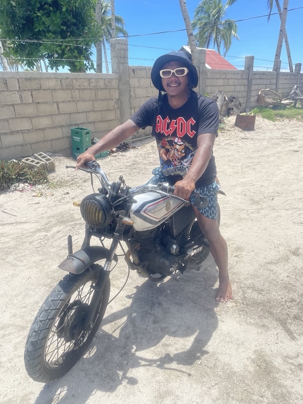 "Ambum sitzt lächelnd auf seinem Motorrad. Er trägt einen Bucket Hat, eine Sonnenbrille mit dickem weißen Rahmen und ein AC-DC T-Shirt."