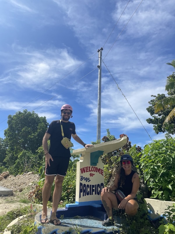 "Sophia und ich posieren vorm Ortseingang von Pacifico: einem steinernen Surfbrett auf einer Platform auf der "Welcome to Barangay Pacifico" steht."