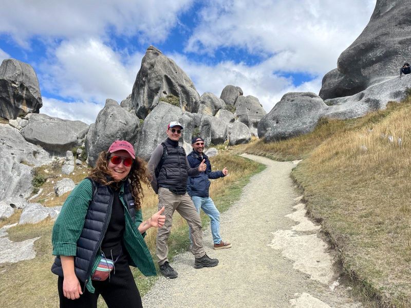 "Sophia, Manu und Simon stehen auf einem Weg mit großen Felsen im Hintergrund unter einem leicht bewölktem Himmel."