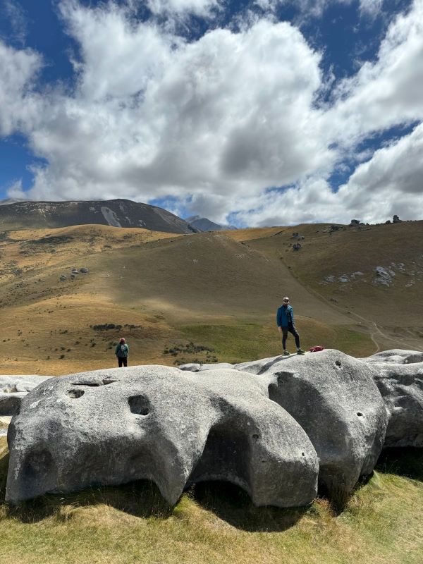 "Sophia und ich stehen in einer felsigen Landschaft voller großer, abgerundeter Felsbrocken im Vordergrund und einer Hügellandschaft im Hintergrund und einer aufgelockerten Bewölkung."