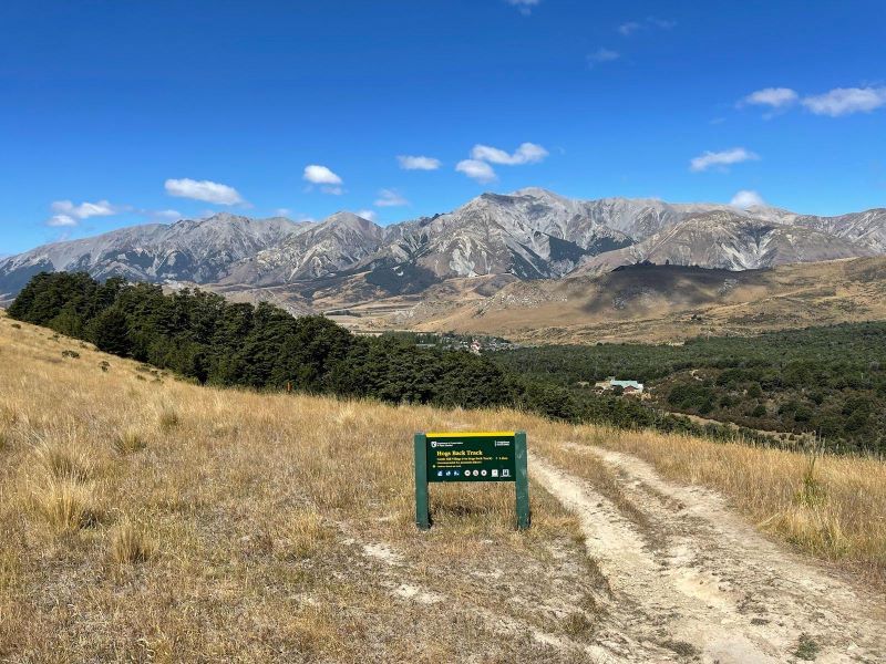 "Eine Berglandschaft mit einem Pfad im Vordergrund, einem Schild dass auf eine Wanderung hinweist und eine Waldlandschaft unter einem blauen Himmel."