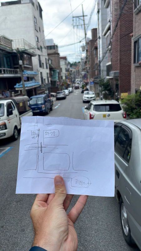 "Ich halte eine handgezeichnete Karte auf einem Blatt Papier hoch. Hinter mir die Straßen von Bomun."