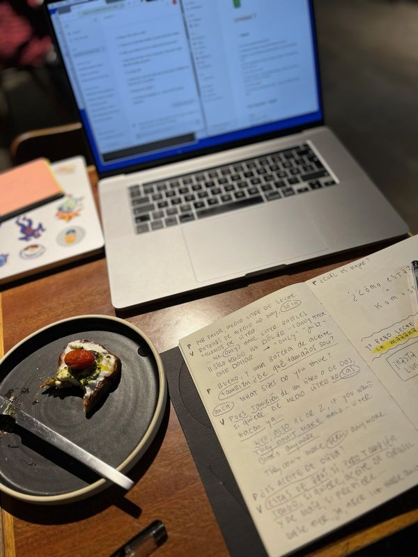 "Am Holztisch des Cafés sind unsere Laptops, ein aufgeschlagenes Notizbuch mit spanischen Vokabeln und ein Teller samt Kuchen."