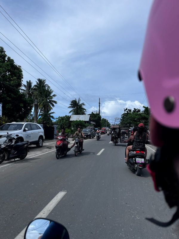 "Ich fahre mit dem Moped auf der STraße. Vor mir ist ein anderes Moped und ein Tuktuk. Einige einzelne Mopedfahrer ohne Helm kommen mir entgegen."