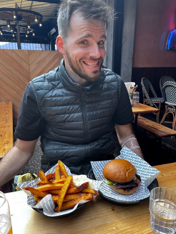 "Ich habe ein schwarzes T-Shirt und eine schwarze Weste an wie ich an einem Tisch sitze. Vor mir sind ein Burger und Pommes, ich grinse."