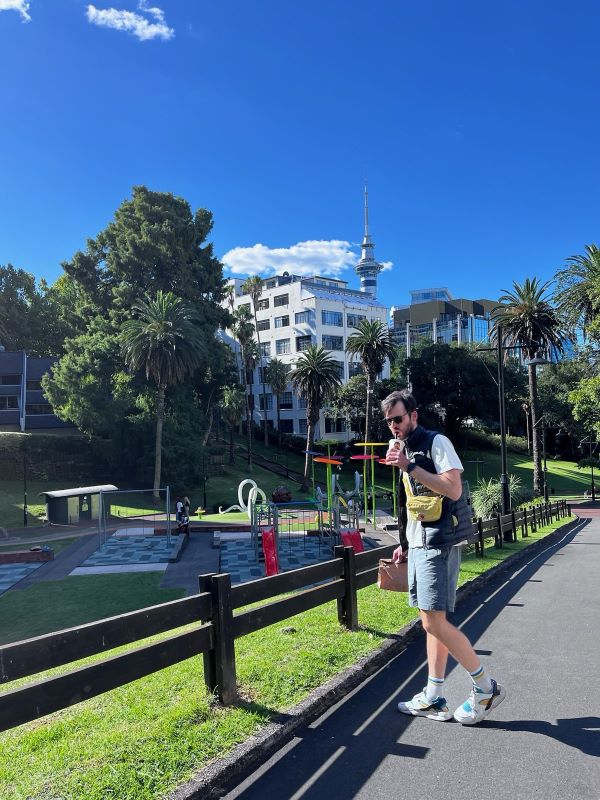 "Ich stehe vor einem Spielplatz im grünen Myers Park, während ich an einem Milkshake schlürfe. Im Hintergrund sind Bäume und der Fernsehturm Aucklands zu sehen."