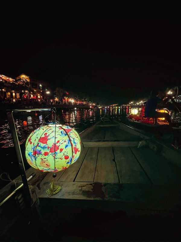 "Auf dem Boot ist eine gelbe Laterne mit blau-gelbem floralen Muster aufgehängt. Am Ufer und am Wasser sind Lichter zu erkennen."